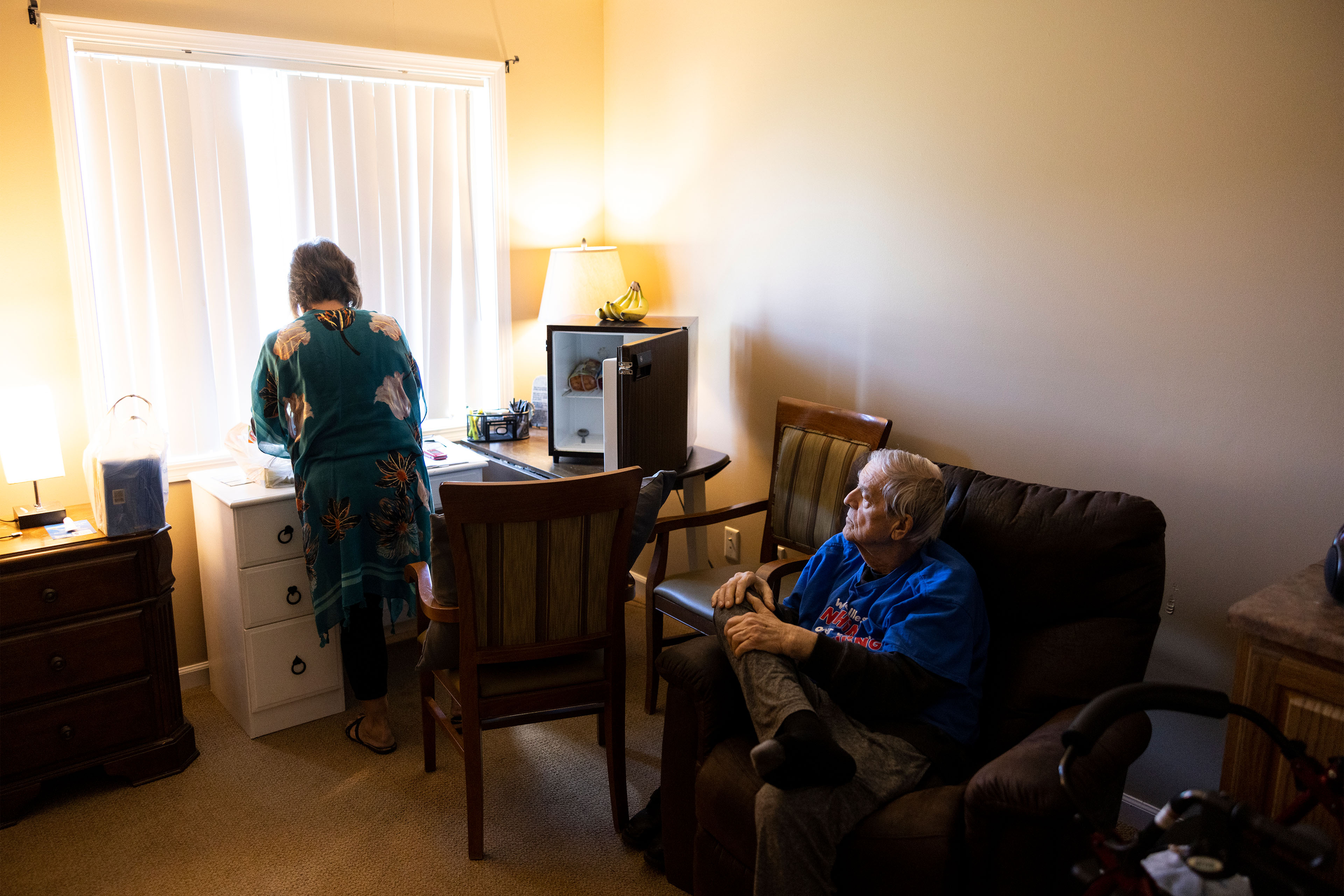 A photo of an elderly man sitting in a chair while a younger woman stands at a nearby drawer with grocery bags.