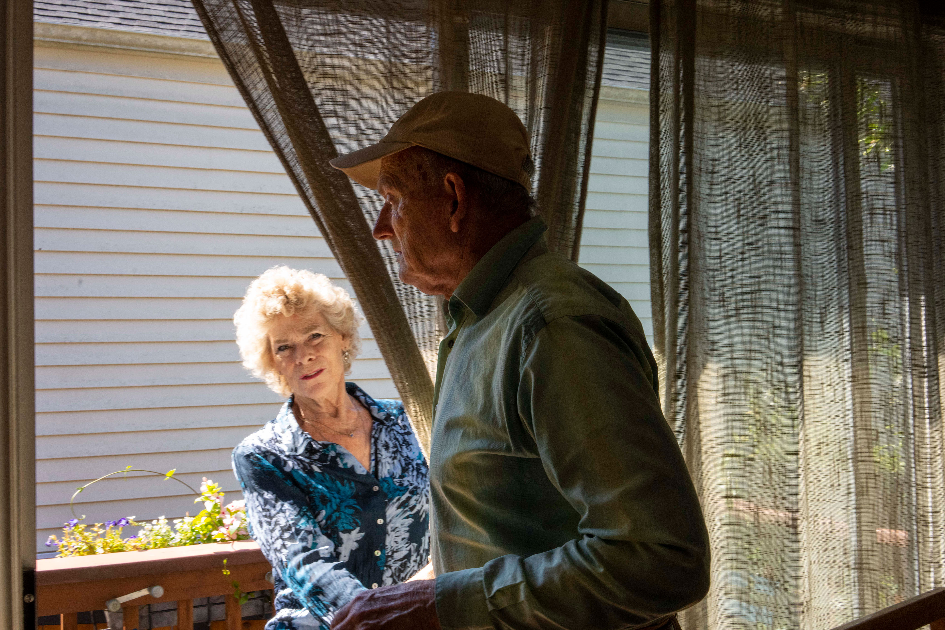 A photo of a woman pulling back curtains while watching her husband, who has dementia.