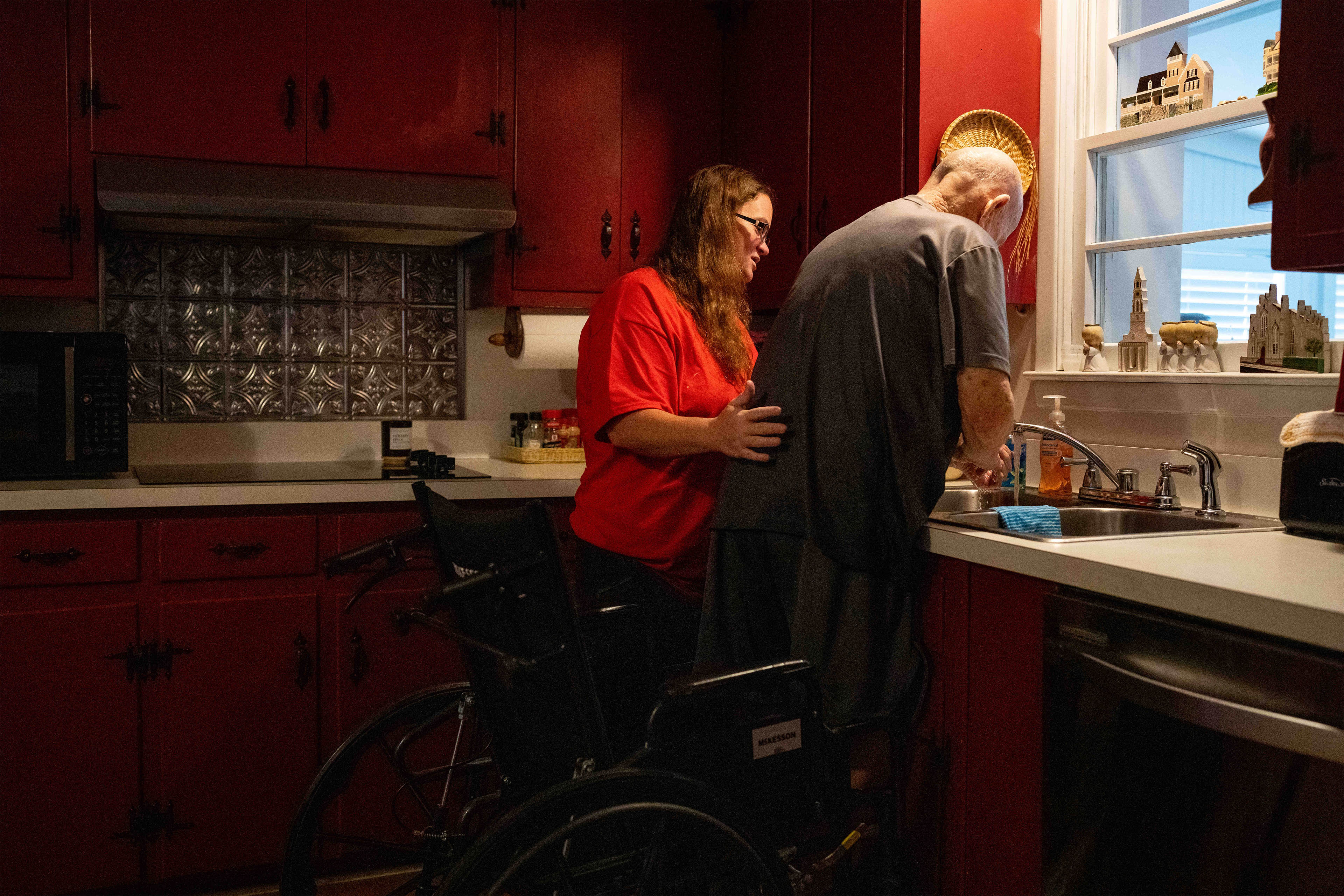 A photo of a caretaker helping an elderly man wash his hands at a kitchen sink.