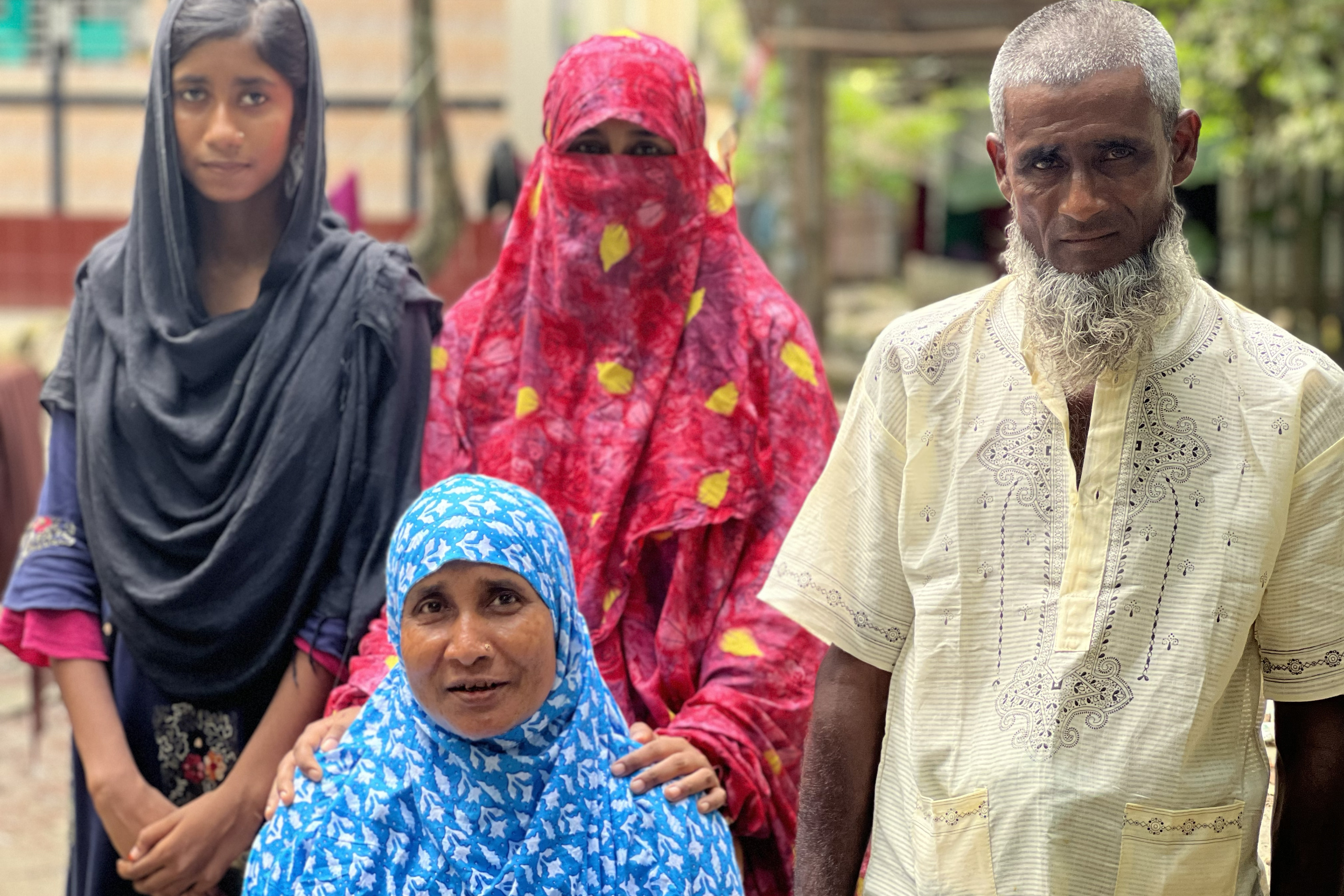 Rahima Banu, seated in front of her husband and two daughters, wears a cobalt-blue scarf with white flowers — it’s draped over her head and shoulders and modestly tucked under her chin. The daughter who stands immediately behind her rests her hands on her mother’s shoulders; she is draped in bright pink cloth that covers all but her eyes. The daughter to the viewer’s left wears a black scarf that half covers her head, and she holds her hands together in front of her. Her husband is in a daffodil-yellow short-sleeved shirt, his gaze fixed on the camera in front of him.