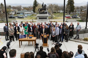 Gov. Greg Gianforte, surrounded by Republican lawmakers, speaks at a bill signing ceremony on the steps of the State Capitol, in Helena, MT. Press huddles in front of the podium he speaks from with microphones and cameras.