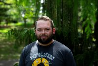 A man with brown hair and bear stands outside by a tree and looks at the camera.