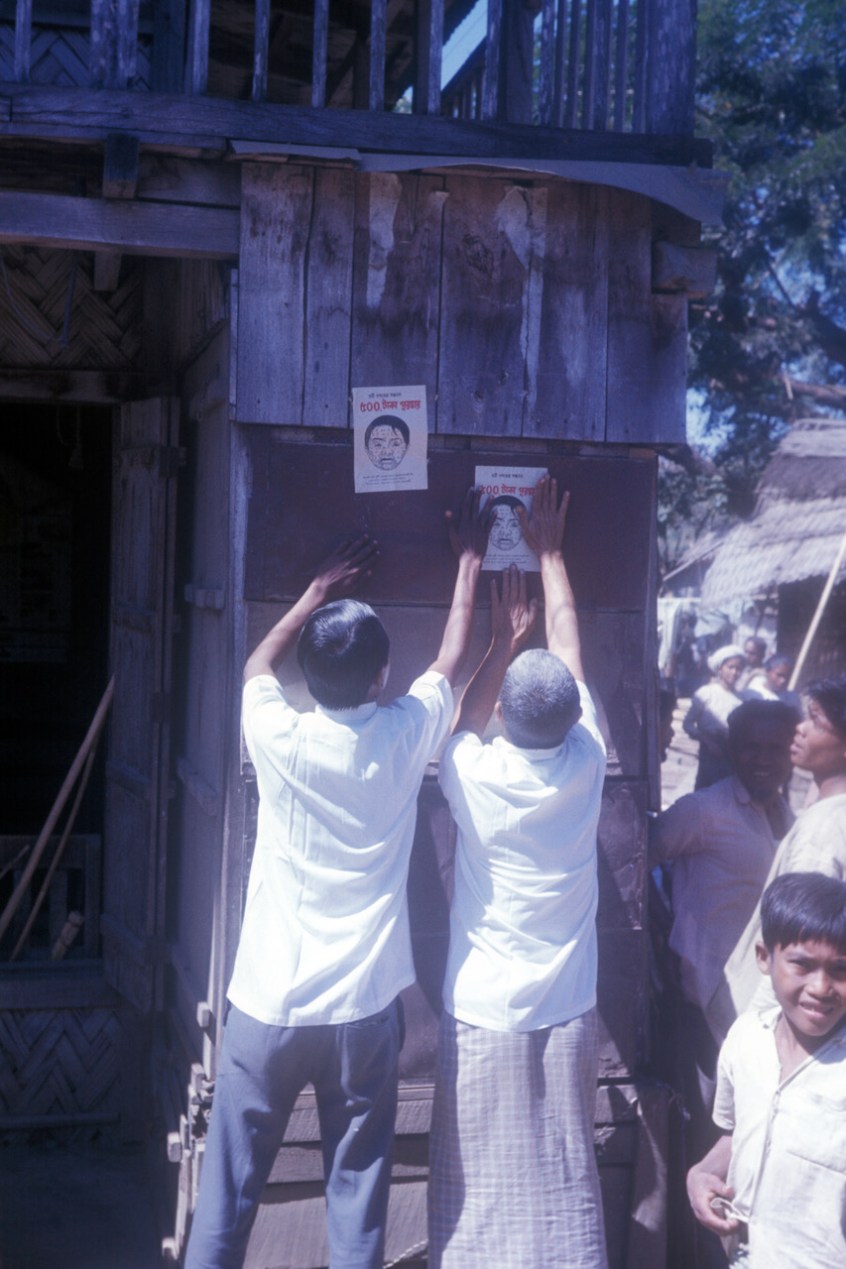 A color film photograph from circa 1975 shows two men pasting up flyers on the side of a wooden building. The flyers show a drawing of a face with smallpox pustules.