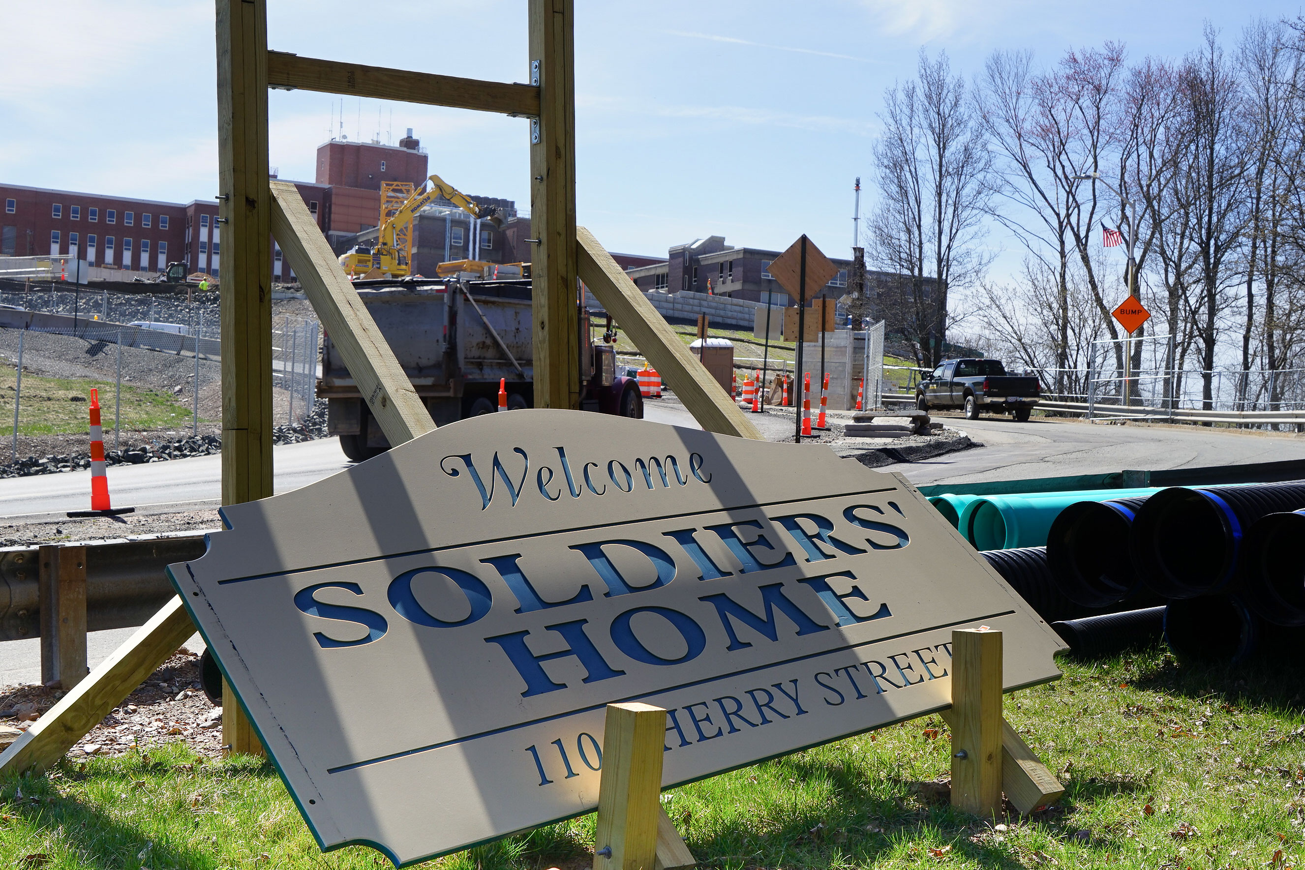 A sign in the midst of being constructed reads "Welcome, Soldiers' Home, 110 Cherry Street." A driveway behind the sign leads up a hill to a large brick building. There is more construction around the driveway in front of the building.