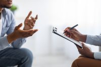 A photo of a man speaking to a therapist across from him, who is writing on a clipboard.