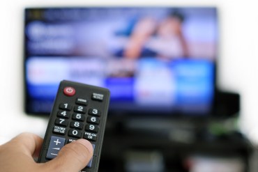 A hand is holding a remote control, there is a blurred TV screen in the background.