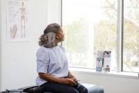 A woman of color sits in a doctor's office. She looks out the window, facing away from the camera.