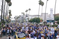 A photo of a crowd of protesters holding signs and marching down a street in Los Angeles. Their signs have slogans such as "Fair wages for healthcare workers," and "Our patients need more staff."