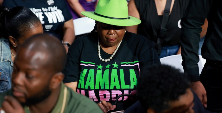 A photo of a Black women wearing a Kamala Harris shirt and a green hat that casts her eyes in shadow.