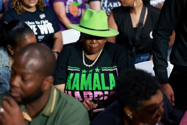 A photo of a Black women wearing a Kamala Harris shirt and a green hat that casts her eyes in shadow.