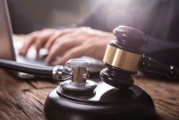 A close-up photograph of a stethoscope and wooden gavel, with a person using a laptop computer in the background.