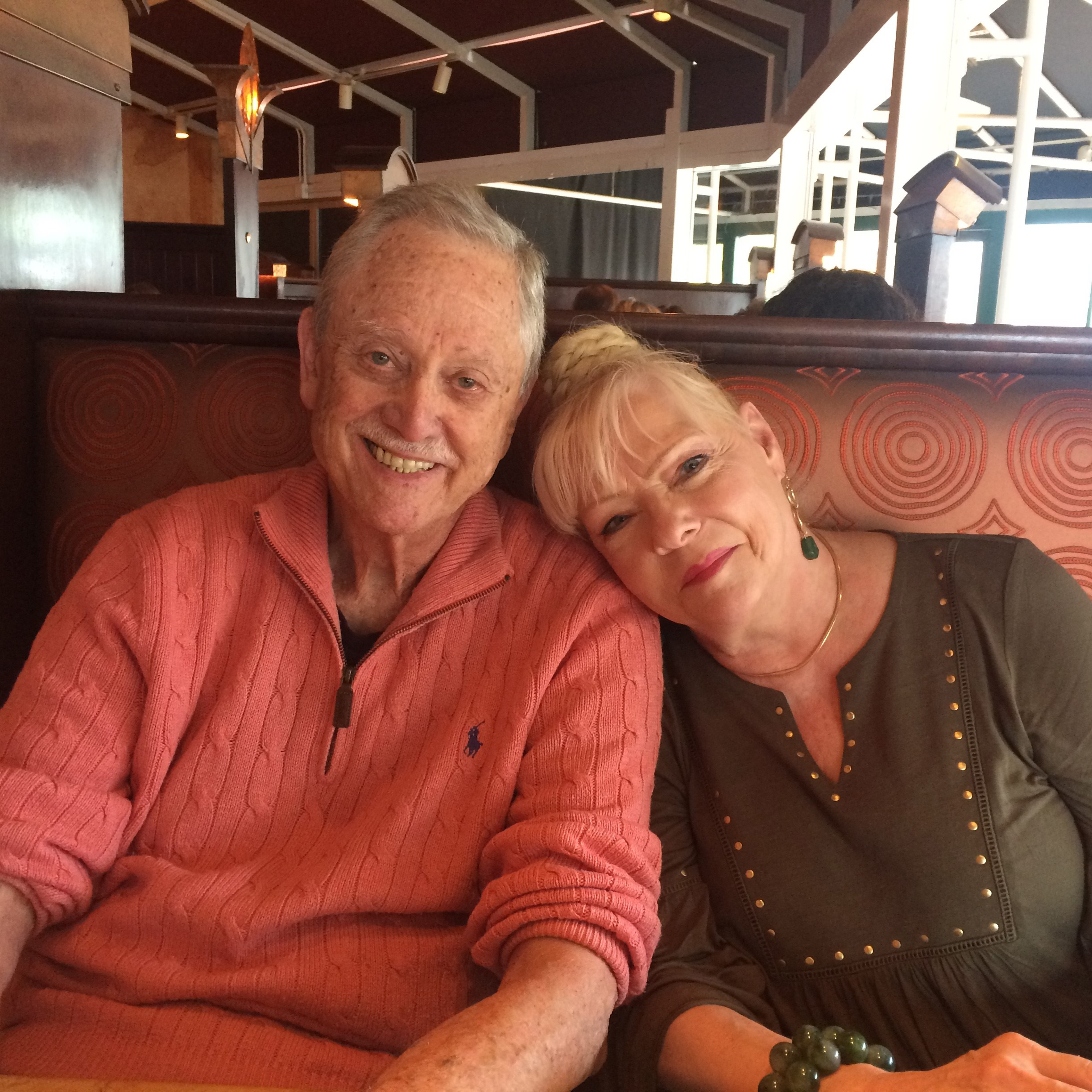 A man in a salmon colored sweater is seated beside a blonde woman, resting her head on his shoulders