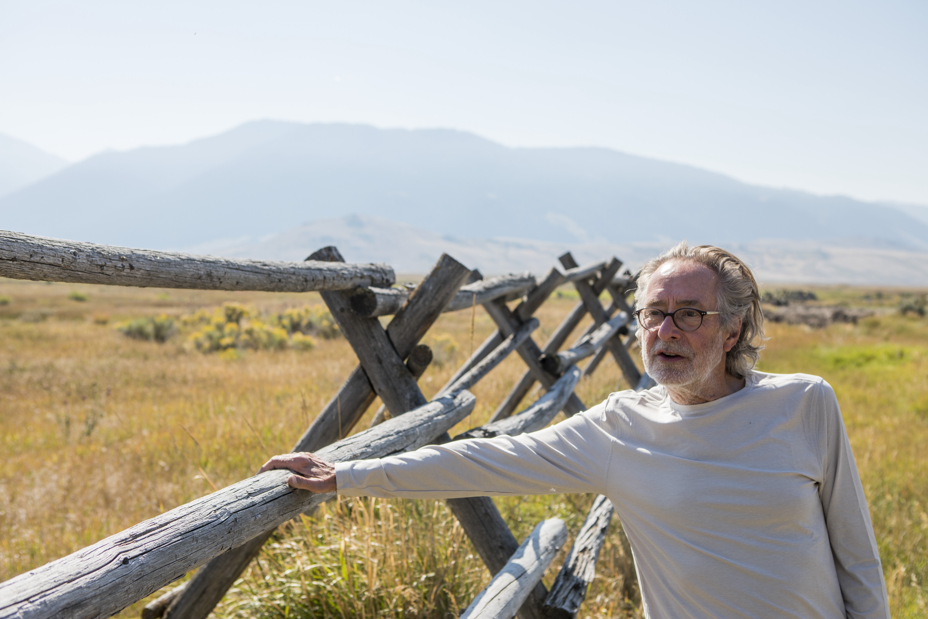 A man in a light colored long sleeve tshirt and glasses stands outside near a wooden fence