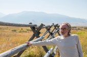 A man in a light colored long sleeve tshirt and glasses stands outside near a wooden fence