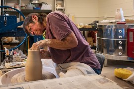 Denise Baker, a senior woman, works at a pottery wheel in a ceramics studio space.