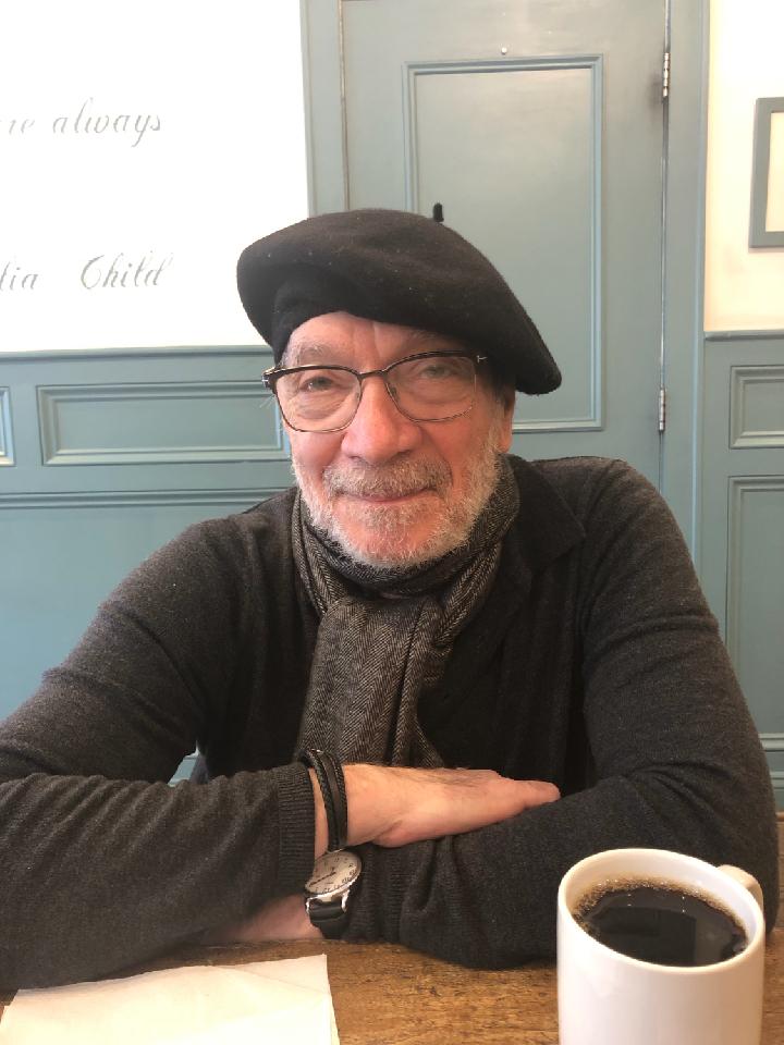 A senior man wearing glasses, a black beret, and a black sweater sits at a table, a cup of black coffee is in front of him