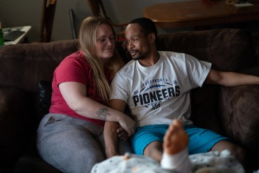 A woman and man sit on a couch, smiling at each other, with arms intertwined