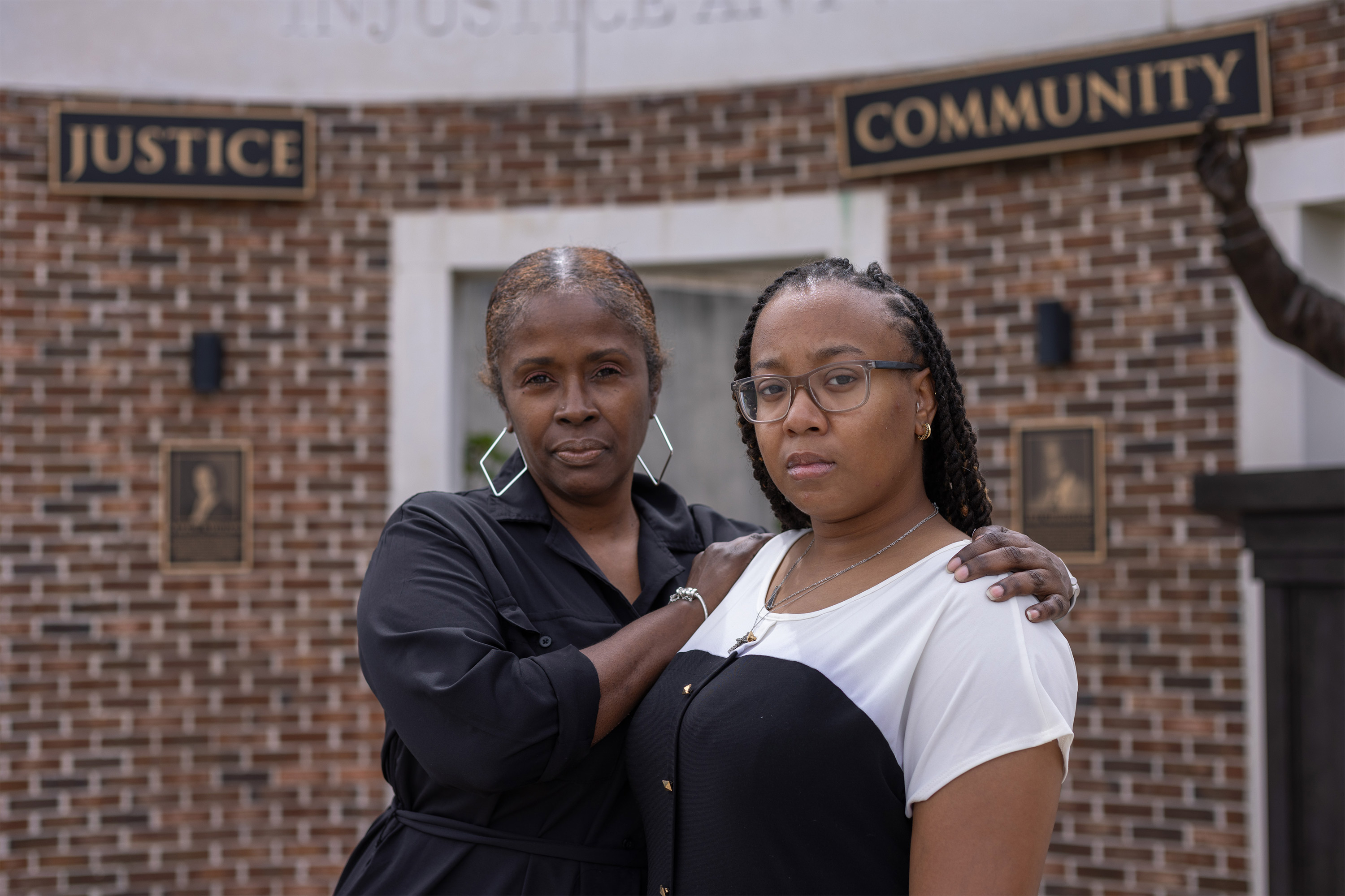 A photo of Zipporah Sumpter with her arms around Amari Marsh's shoulders. Both are looking at the camera. They are standing by a structure with plaques behind them that read, "Justice" and "Community."