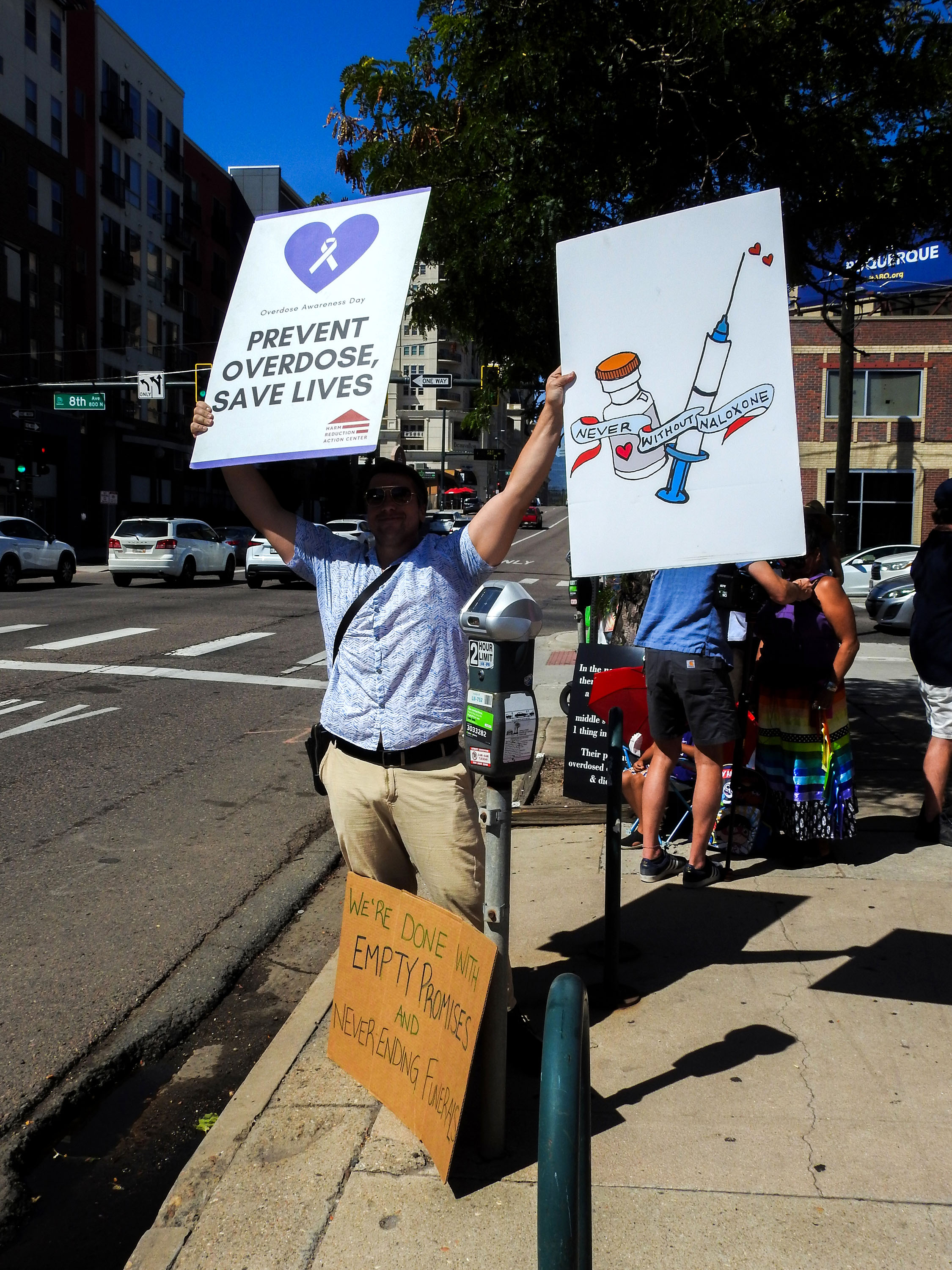 A man is standing on the side of a main road holding up two signs. The sign on the left says: "Prevent overdose, save lives." The sign on the right shows a drawing of a syringe and vial, with words that say: Never without Naloxone." A third sign is leaning up agains the man's legs. It reads: "We're done with empty promises and never ending funerals."