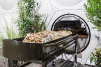 A photo shows a demonstration of a human composting vessel, a horizontal chamber filled with wood chips and other biodegradable materials.