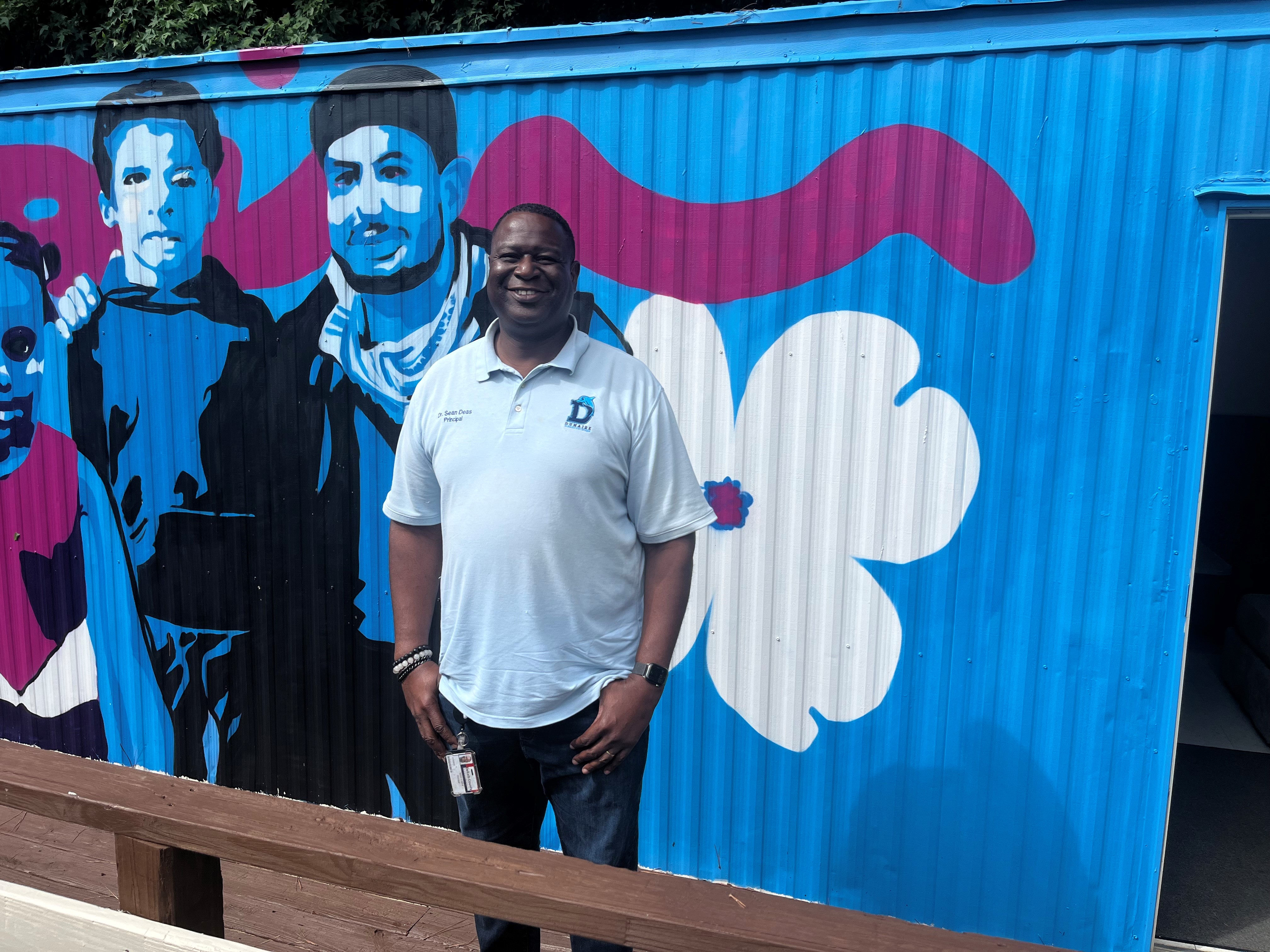 A man in a polo shirt stands in front of a mural with a large white flower and a portrait of three young people