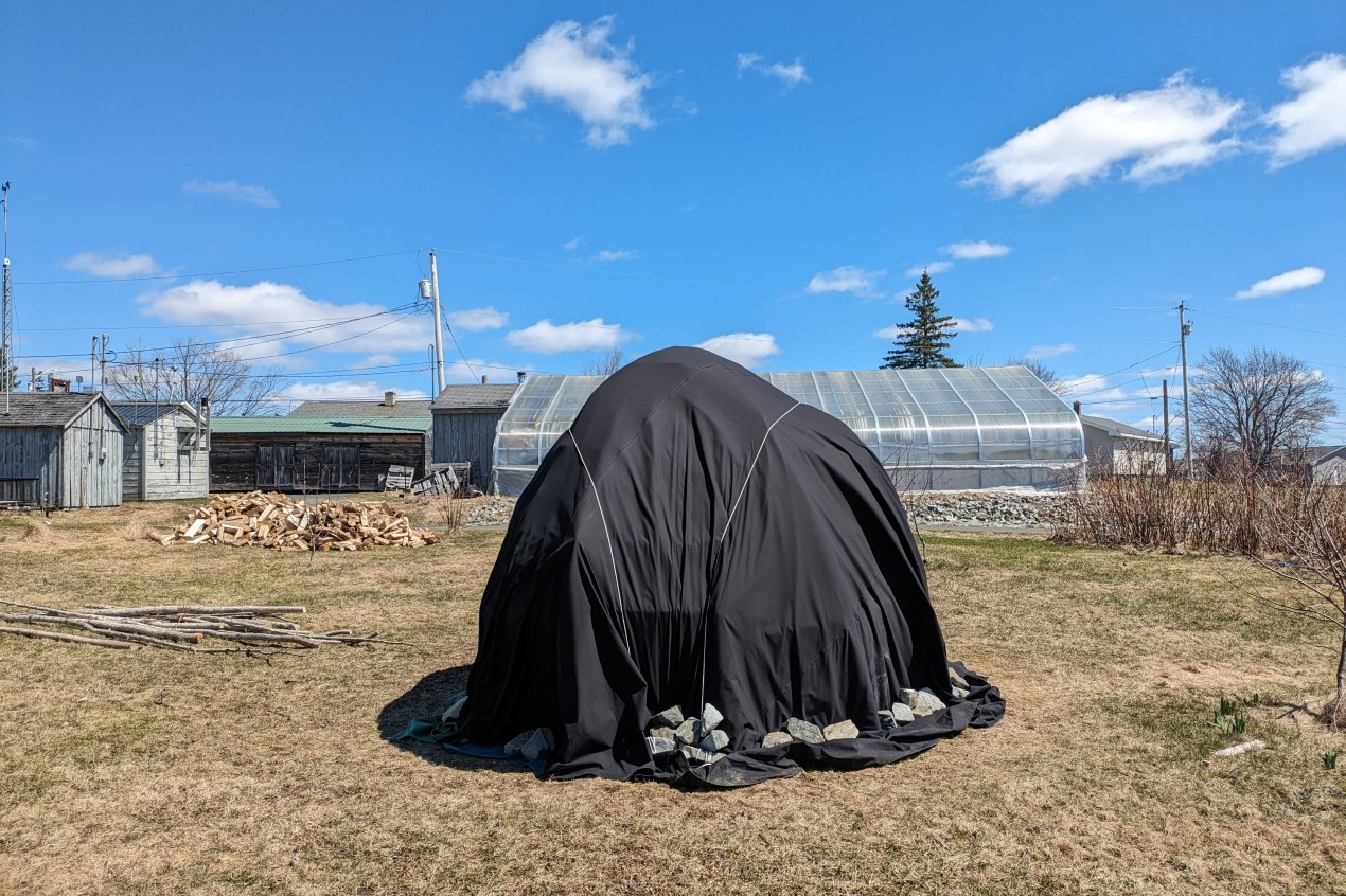 A structure in a clearing between buildings is covered in black fabric weighted by large rocks at the bottom.